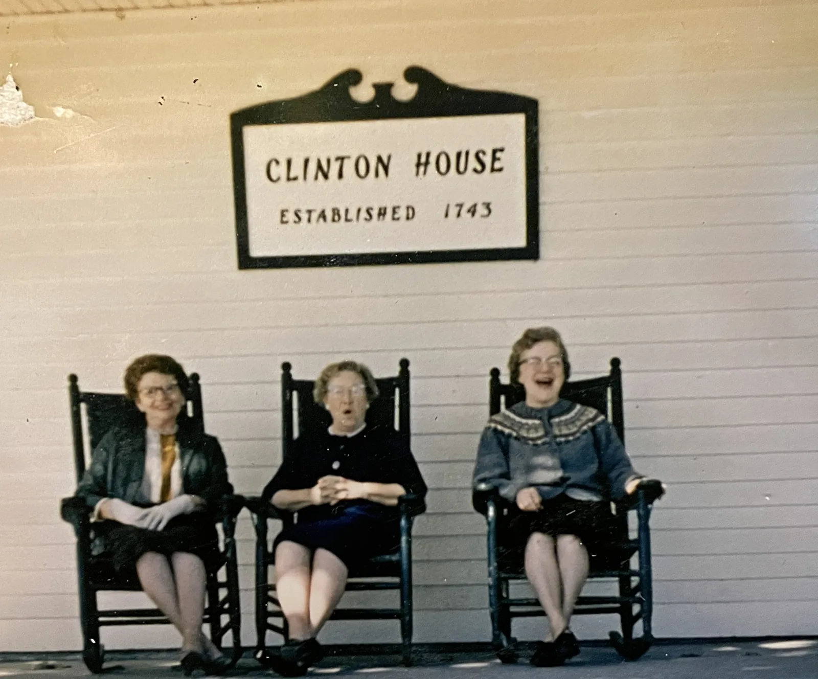 Three women laughing in rocking chairs beneath the carved Clinton House sign reading Established 1743